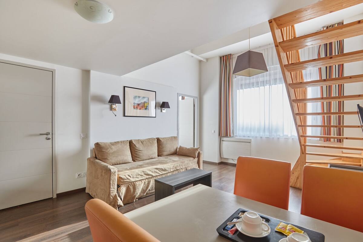Ein modernes Hotelzimmer mit Sofa, Esstisch, Stühlen, Bücherregal und einem fensterbepflanzten Fenster.