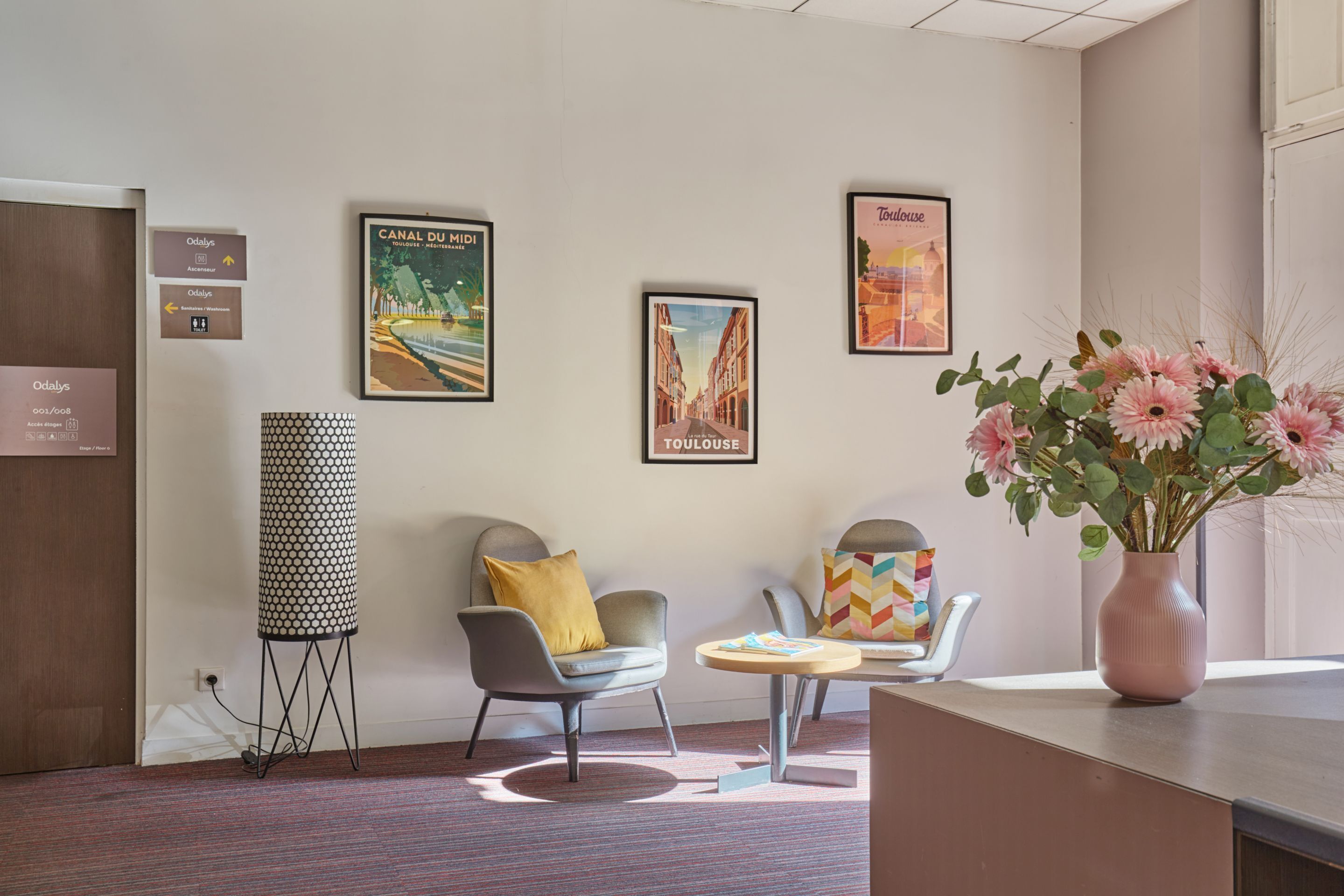 A hotel lobby with a reception desk, a patterned floor lamp, a chair with a yellow pillow, a small round table, and a vase with flowers.