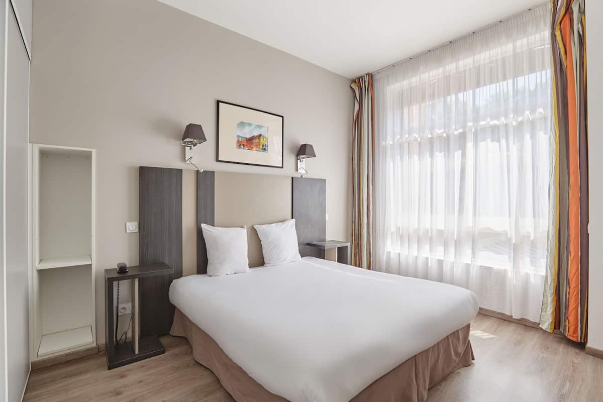 Ein Hotelzimmer mit Bett, Nachttischen, einem Wandbild in einem Rahmen und durchsichtigen Vorhängen.