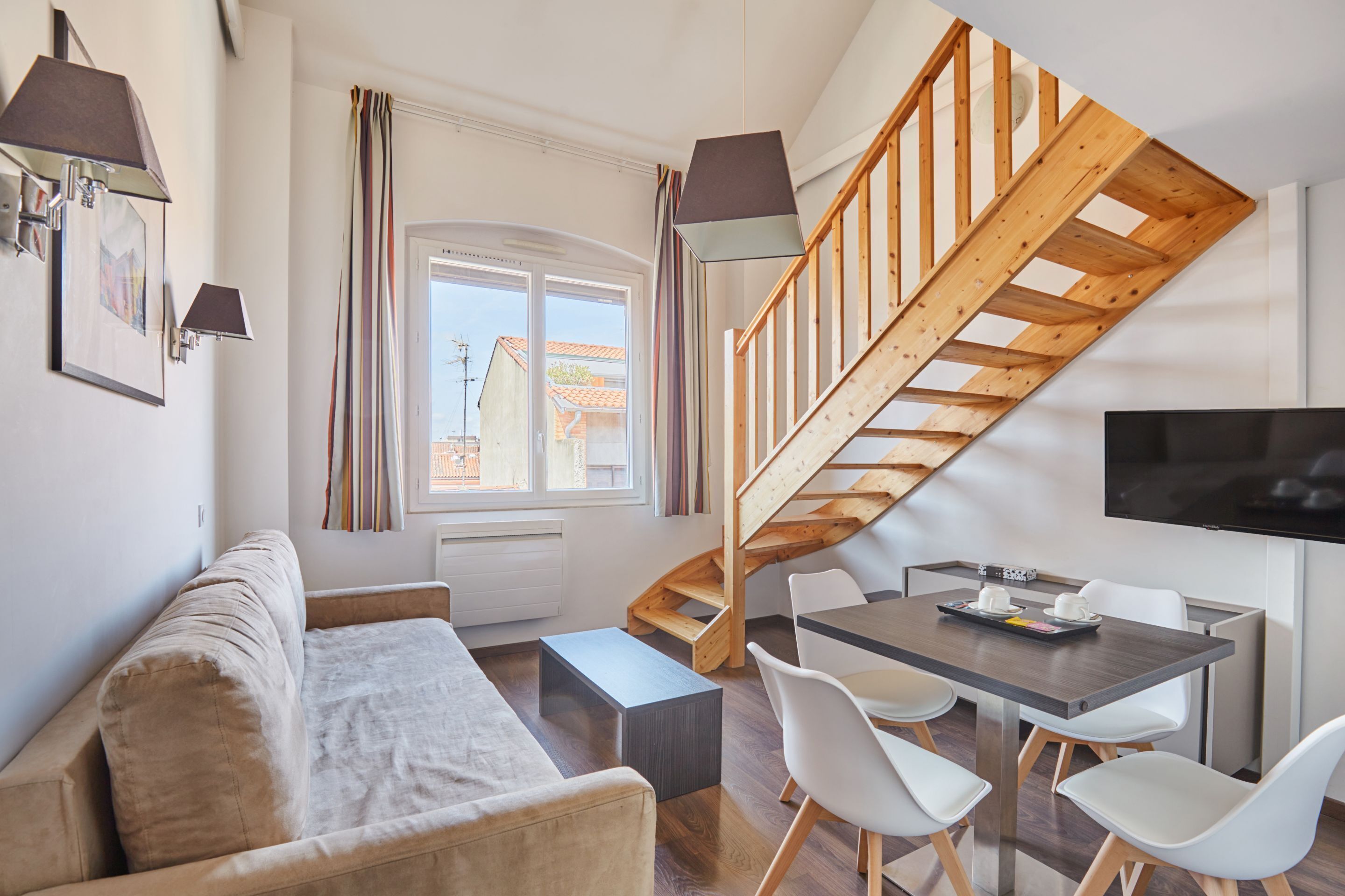 Ein zeitgemäßer Wohnraum mit Sofa, Esstisch, Stühlen, einer Treppe und einem Fernseher.