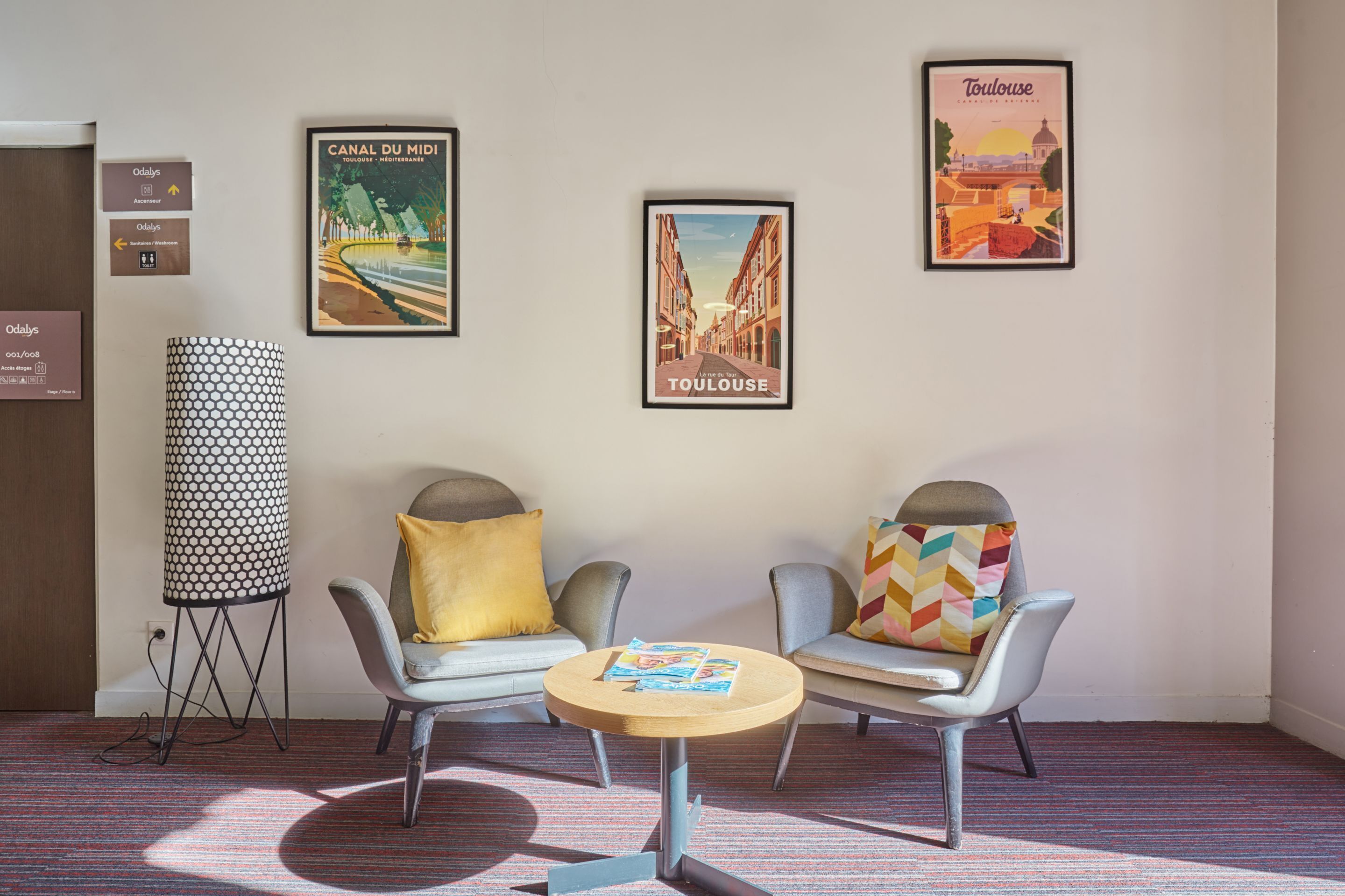 A hotel lobby with two armchairs, a round table, and three framed posters on the wall.