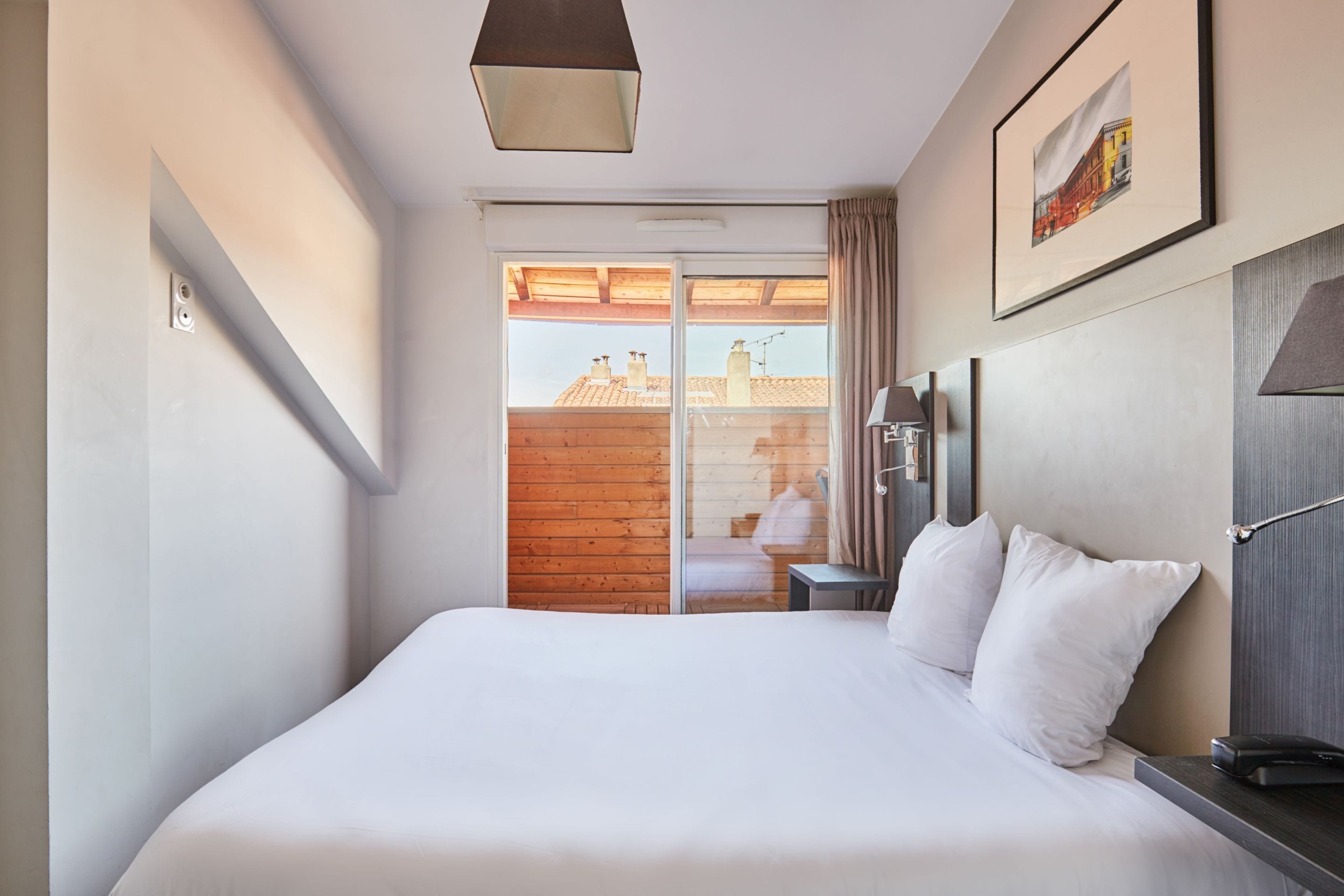 Una habitación de hotel con cama, mesita de noche, lámpara, un cuadro enmarco y vistas al exterior.
