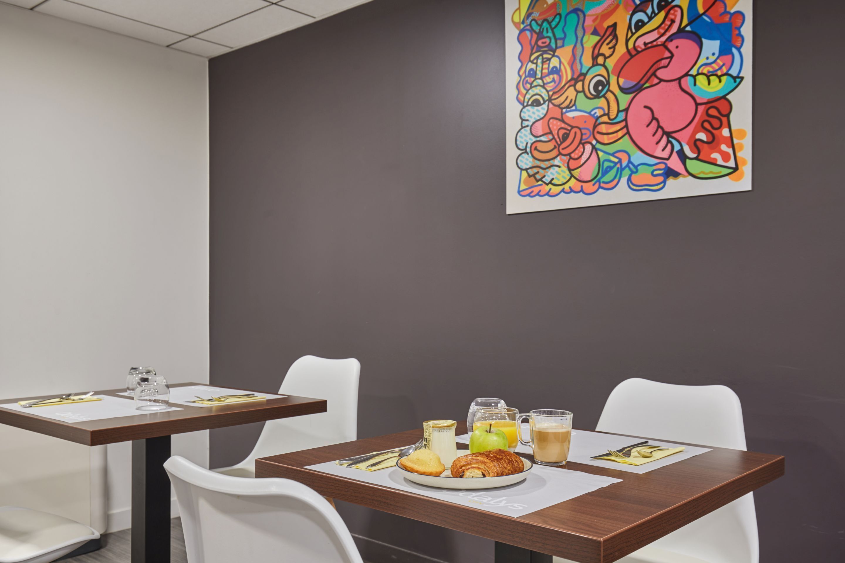A dining area with tables, chairs, and breakfast items including a croissant, apple, orange juice, and coffee.