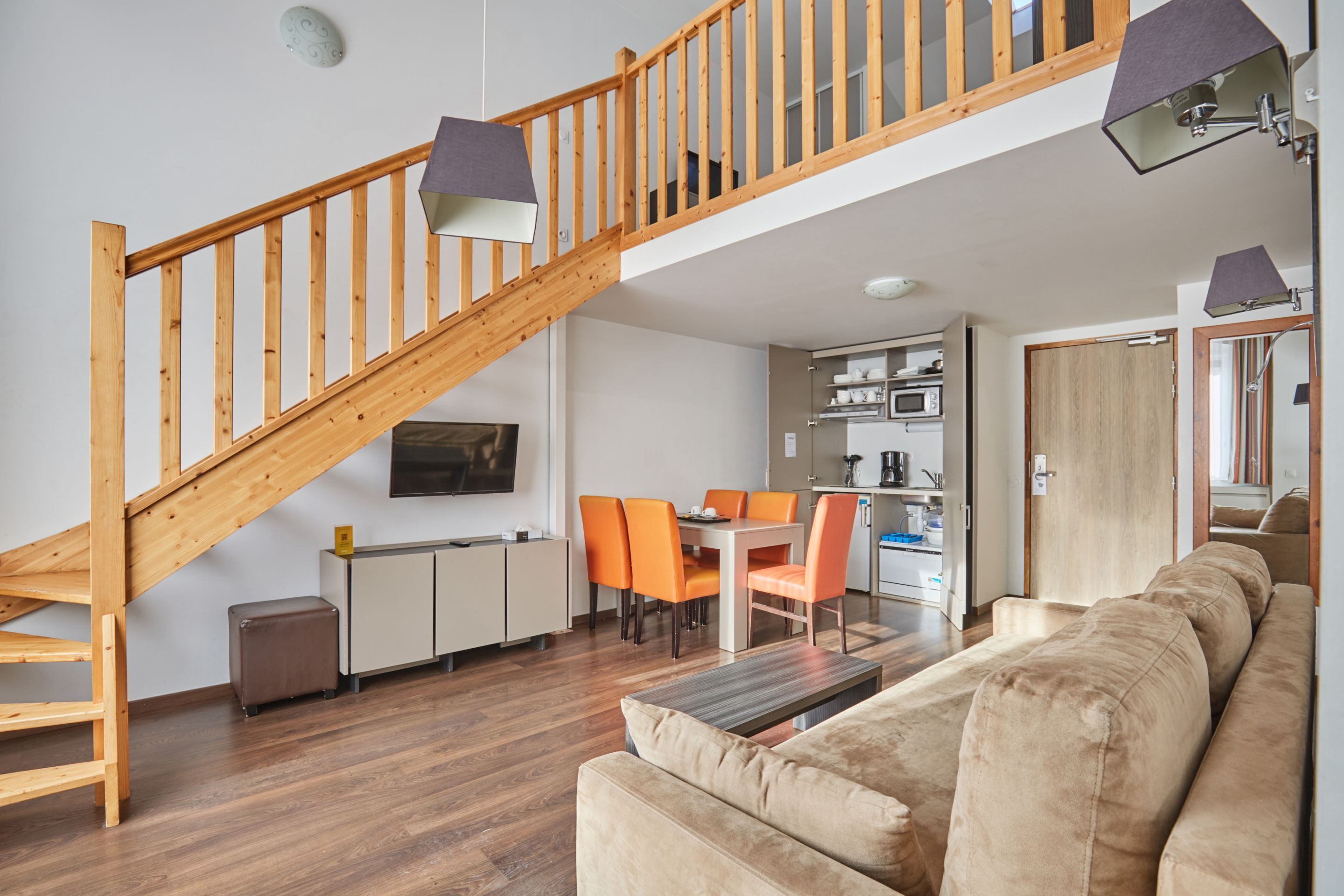 Un intérieur moderne avec un escalier en bois, un coin repas avec des chaises orange, un canapé et une cuisine équipée d’armoires et d’appareils électroménagers.