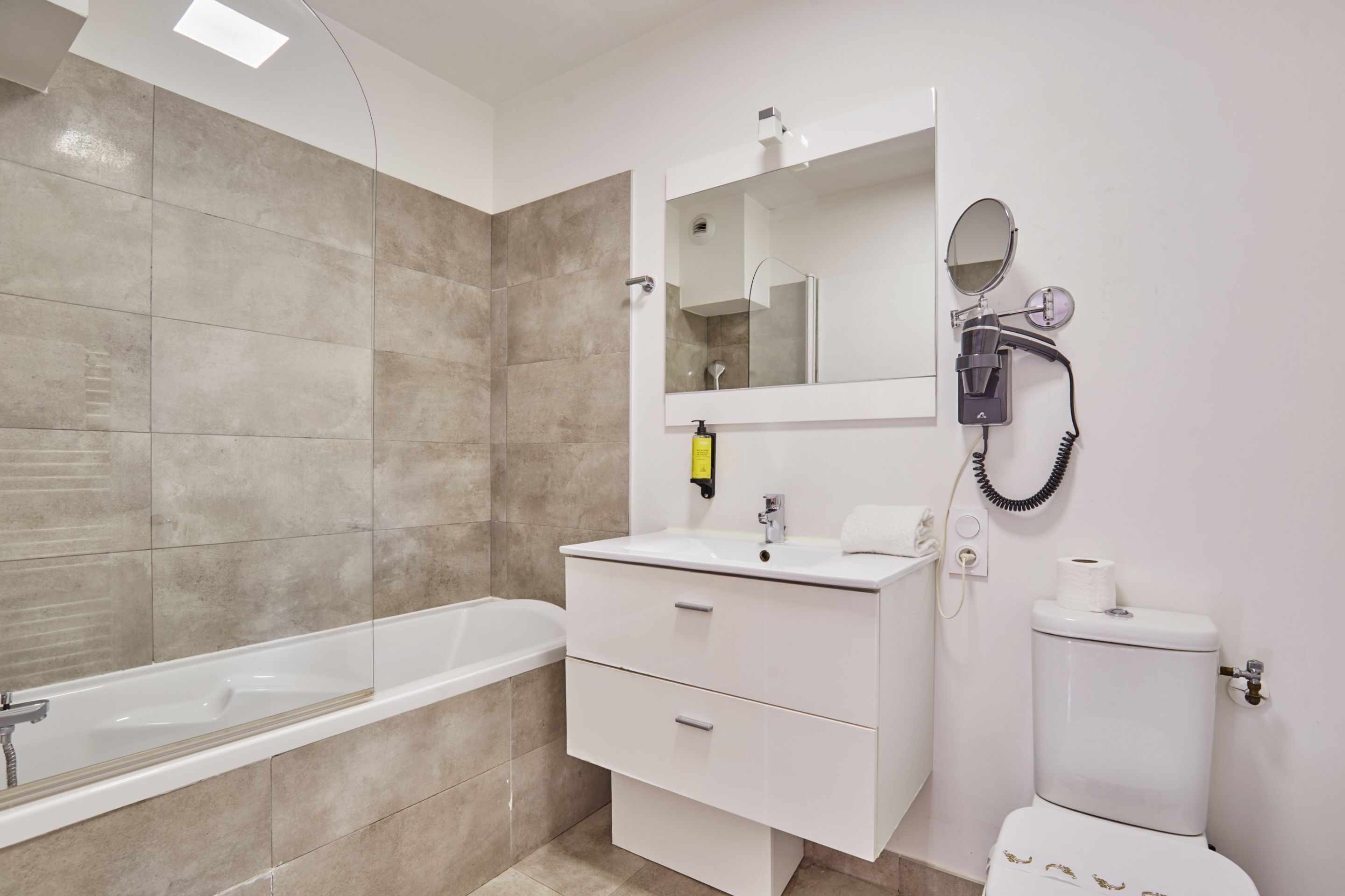A modern bathroom with a bathtub, a white vanity with a mirror, a toilet, and a hairdryer on the wall.