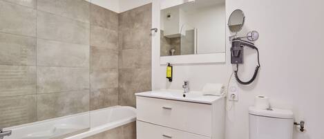 A modern bathroom with a bathtub, a white vanity with a mirror, a toilet, and a hairdryer on the wall.