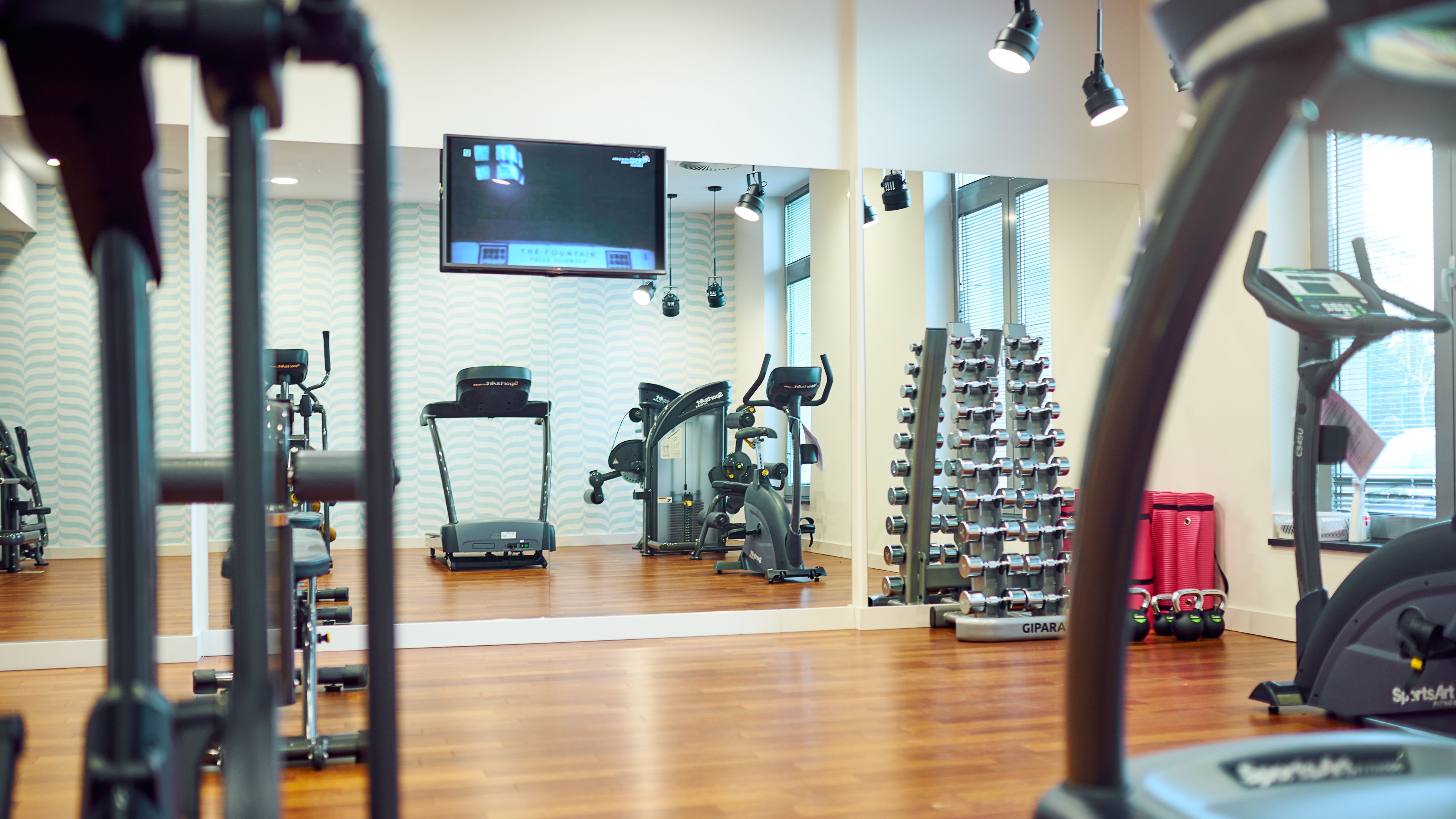 A gym with various exercise equipment, a TV mounted on the wall, and a window with blinds.