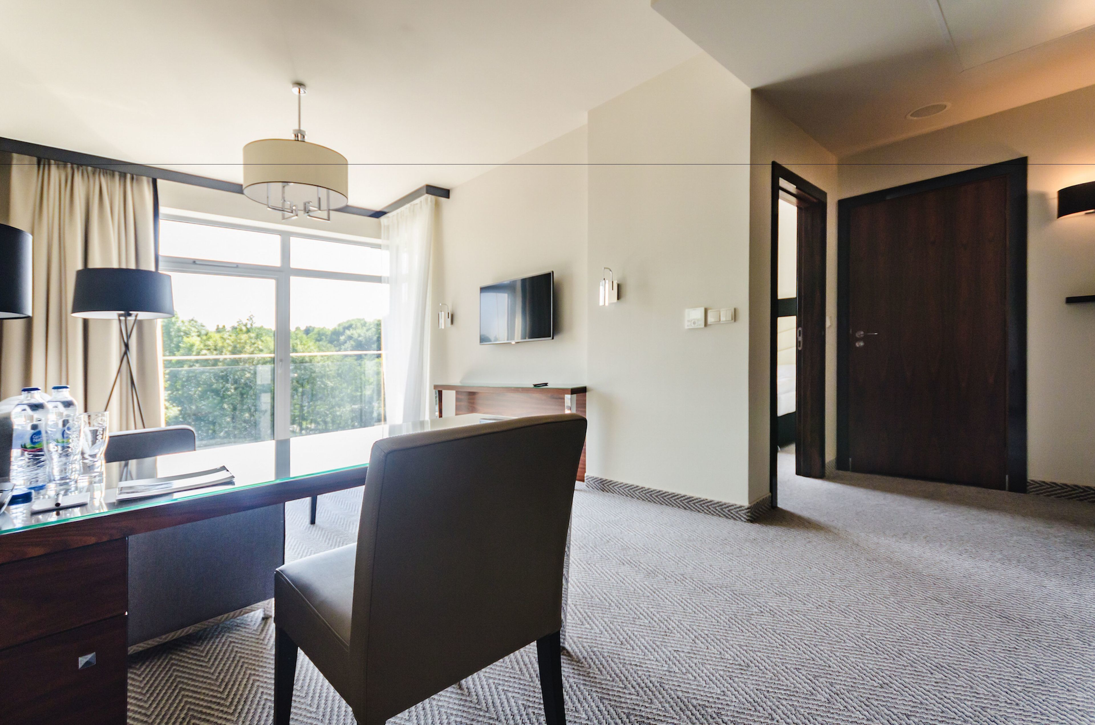 Ein Hotelzimmer mit Schreibtisch, Stuhl, großem Fenster und Fernseher an der Wand.