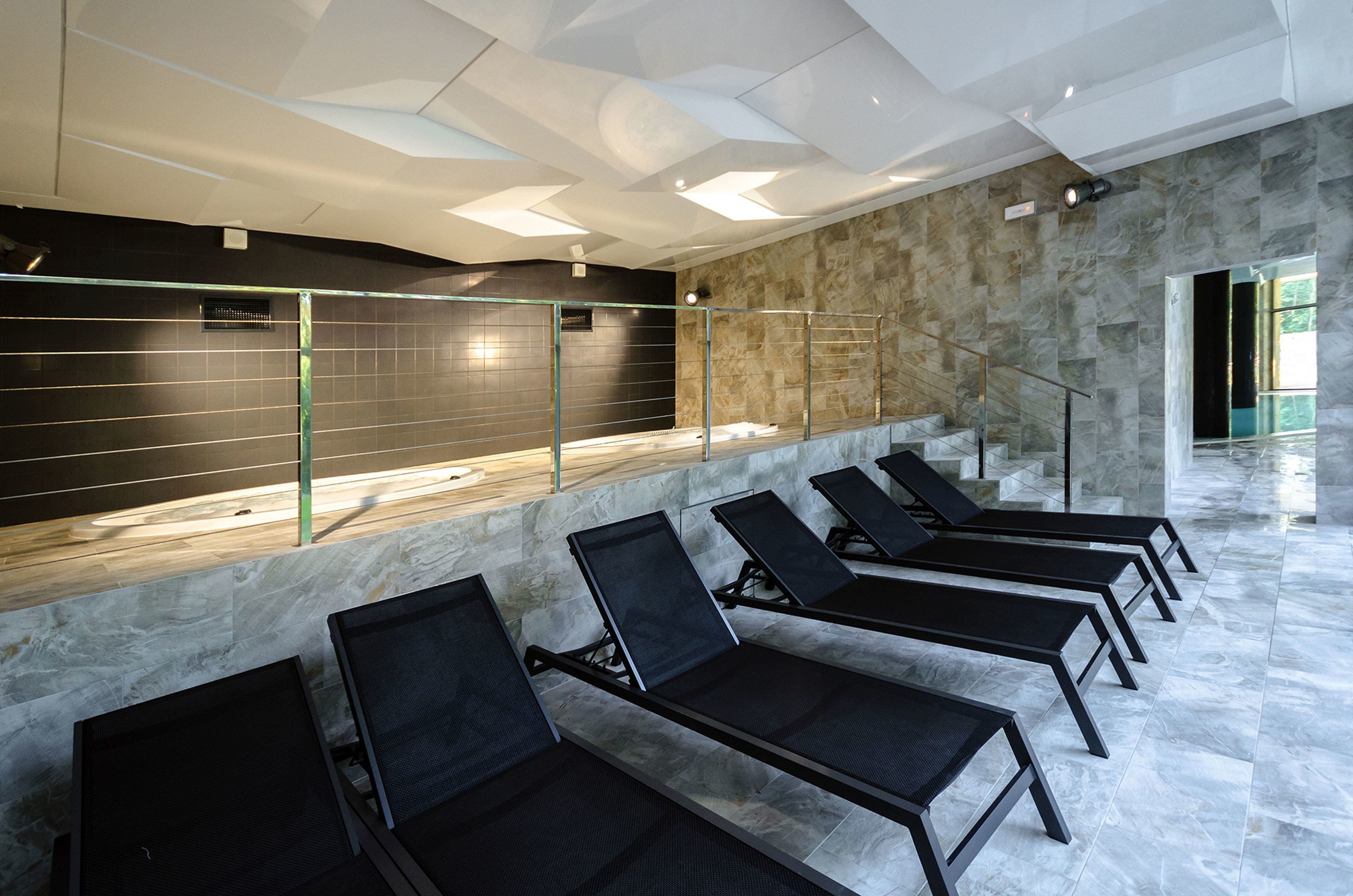 Un moderno espacio de spa con sillones de relax negros, una pared de mármol y una barandilla de vidrio.