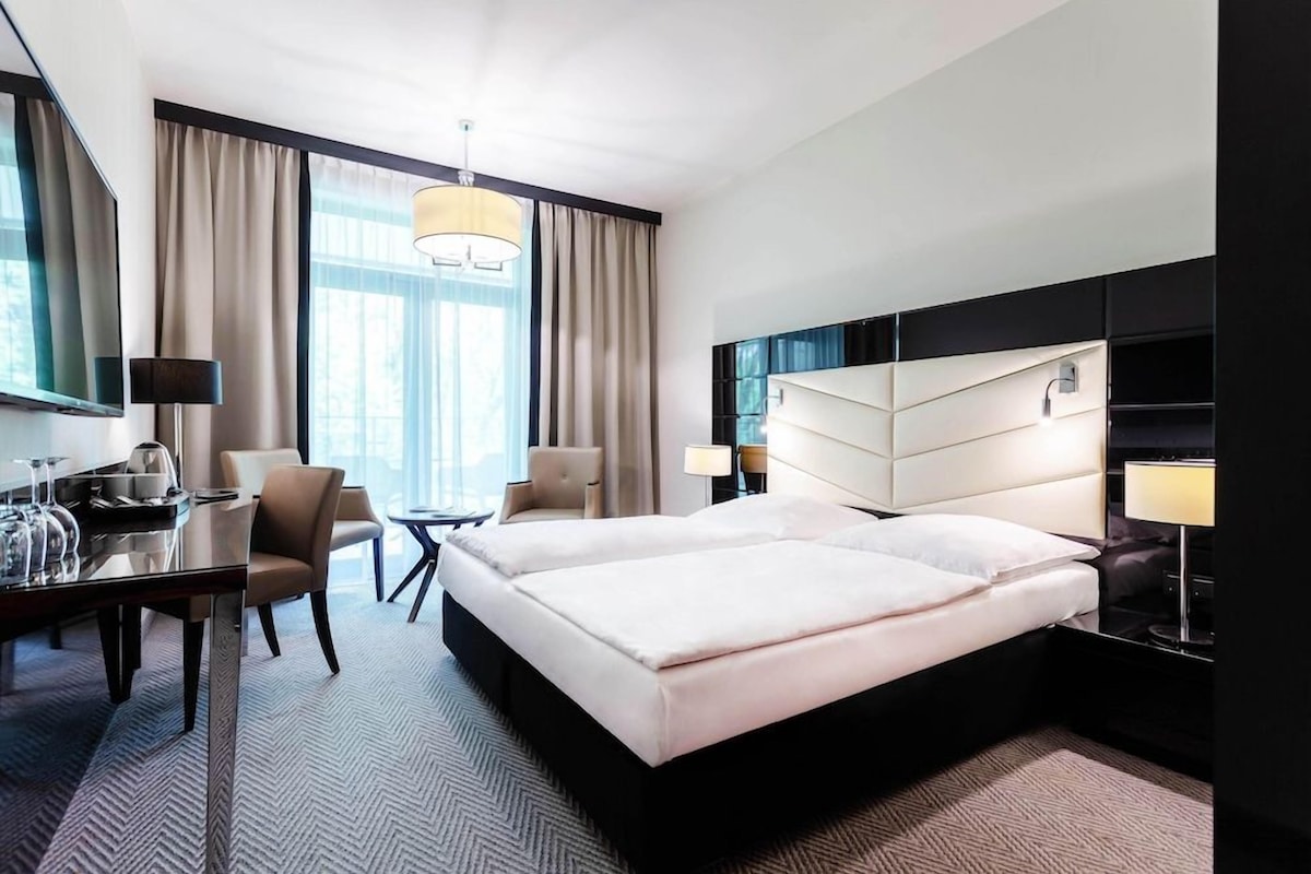 A hotel room with a bed, a desk, a chair, a lamp, and a large window.