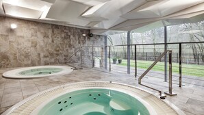 A modern indoor pool area with two hot tubs, a stone wall, and a view of a garden through large windows. - Diune Resort (Kolobrzeg)