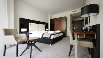 A modern hotel room with a bed, two chairs, a small table, a desk, and a lamp.