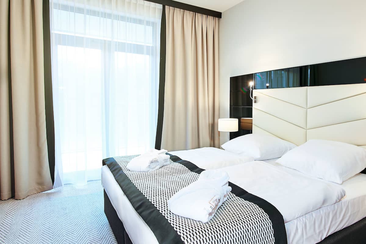 Une chambre d’hôtel avec un grand lit, une tête de lit, une table de chevet, une lampe et une fenêtre avec des rideaux.