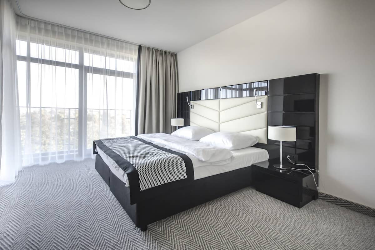 Une chambre d’hôtel moderne avec un grand lit, une table de chevet noire, une petite table de nuit et une vue sur la ville grâce à de grandes fenêtres.