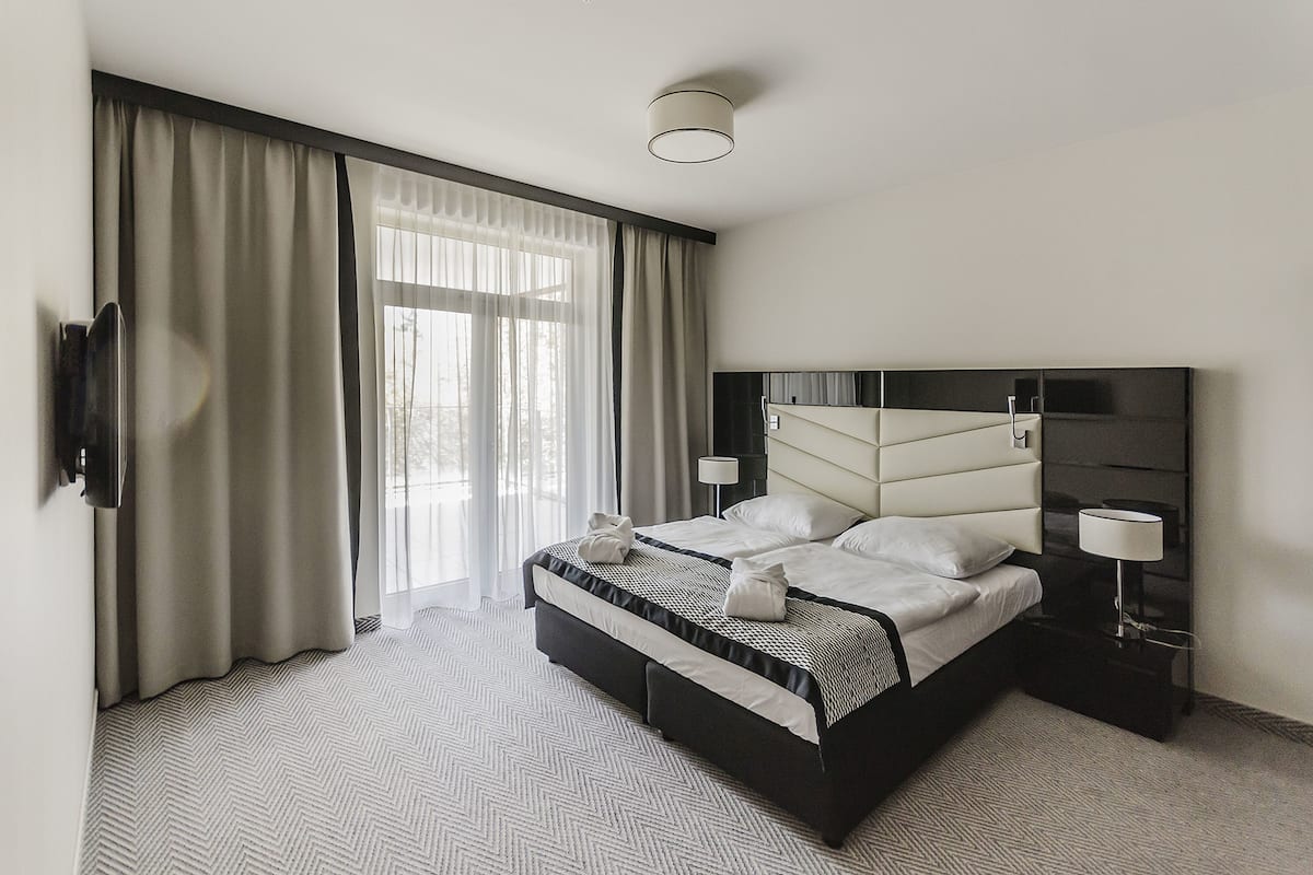 Une chambre d’hôtel moderne dotée d’un grand lit, d’un téléviseur à écran plat, d’une table de chevet avec une lampe et d’une grande fenêtre avec des rideaux.
