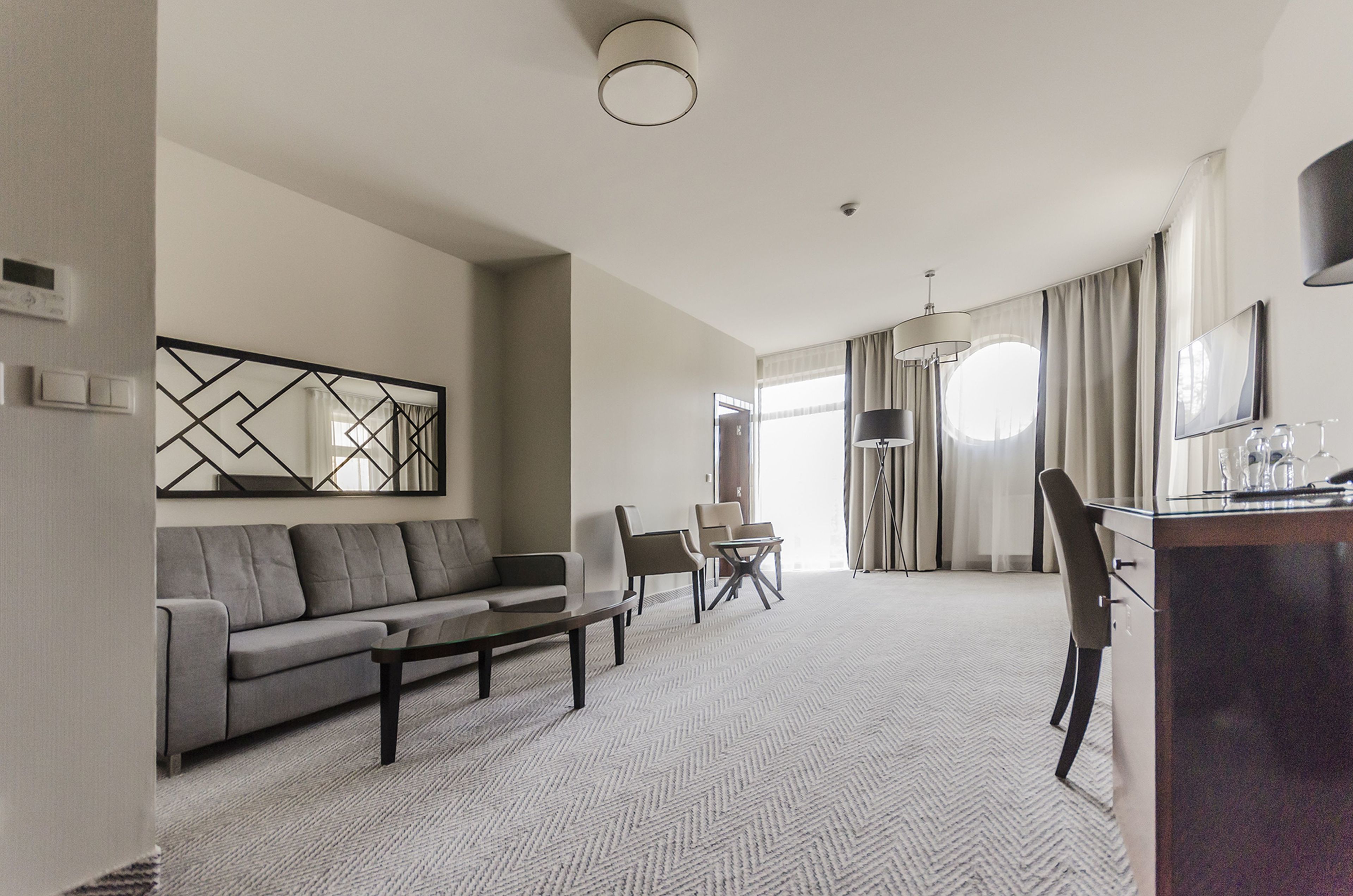 Una habitación de hotel moderna con una sala de estar que incluye un sofá, sillas, una mesa de centro y una mesa de comedor. Hay un escritorio con una lámpara y un televisor. La habitación tiene un ventanal con cortinas y una lámpara de techo.