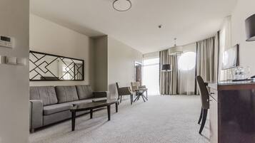 A modern hotel room with a living area featuring a sofa, chairs, a coffee table, and a dining table. There is a desk with a lamp and a TV. The room has a large window with curtains and a ceiling light.