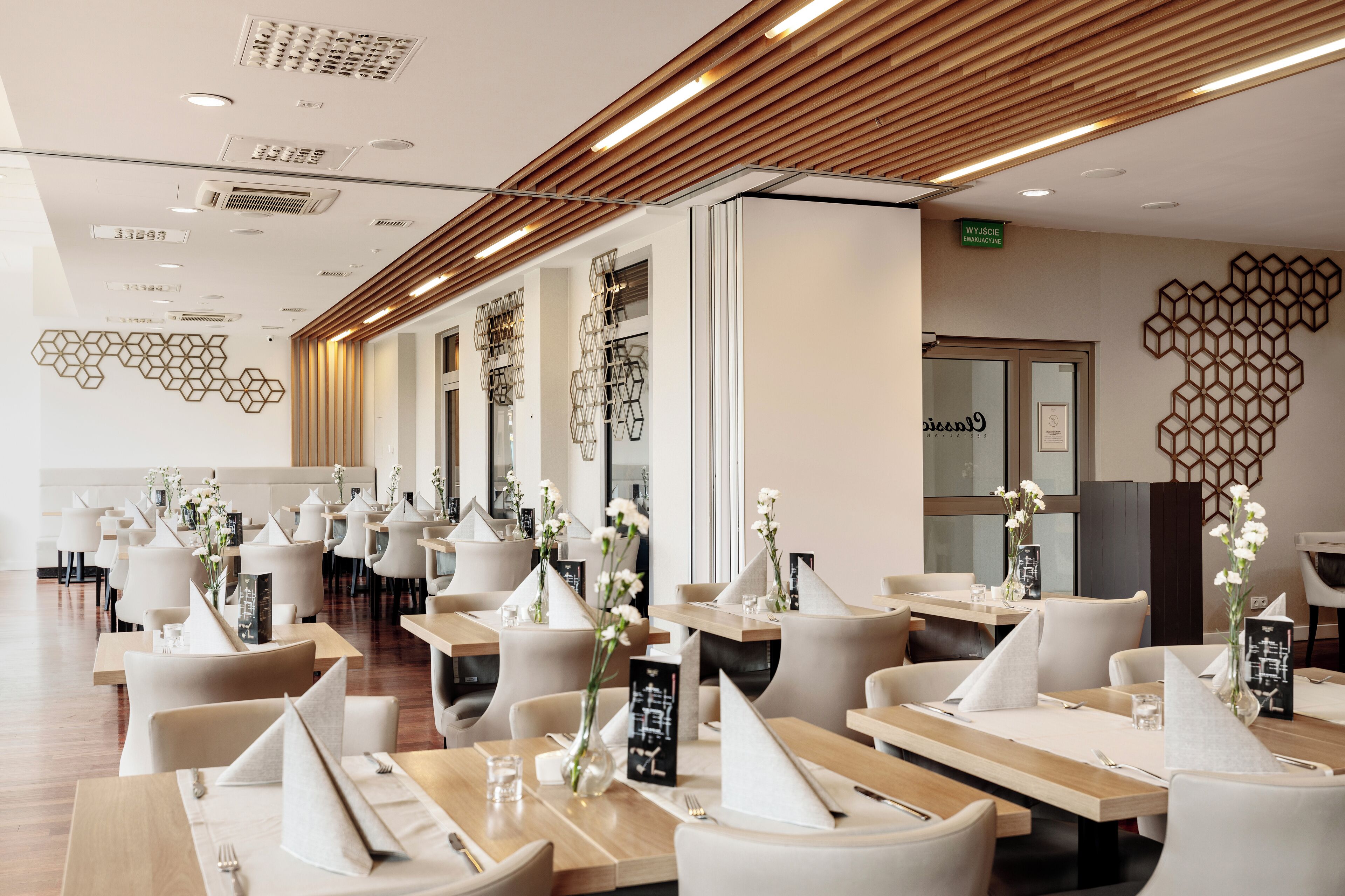 A modern restaurant with multiple tables, each set with white napkins and cutlery, and decorative vases with flowers.