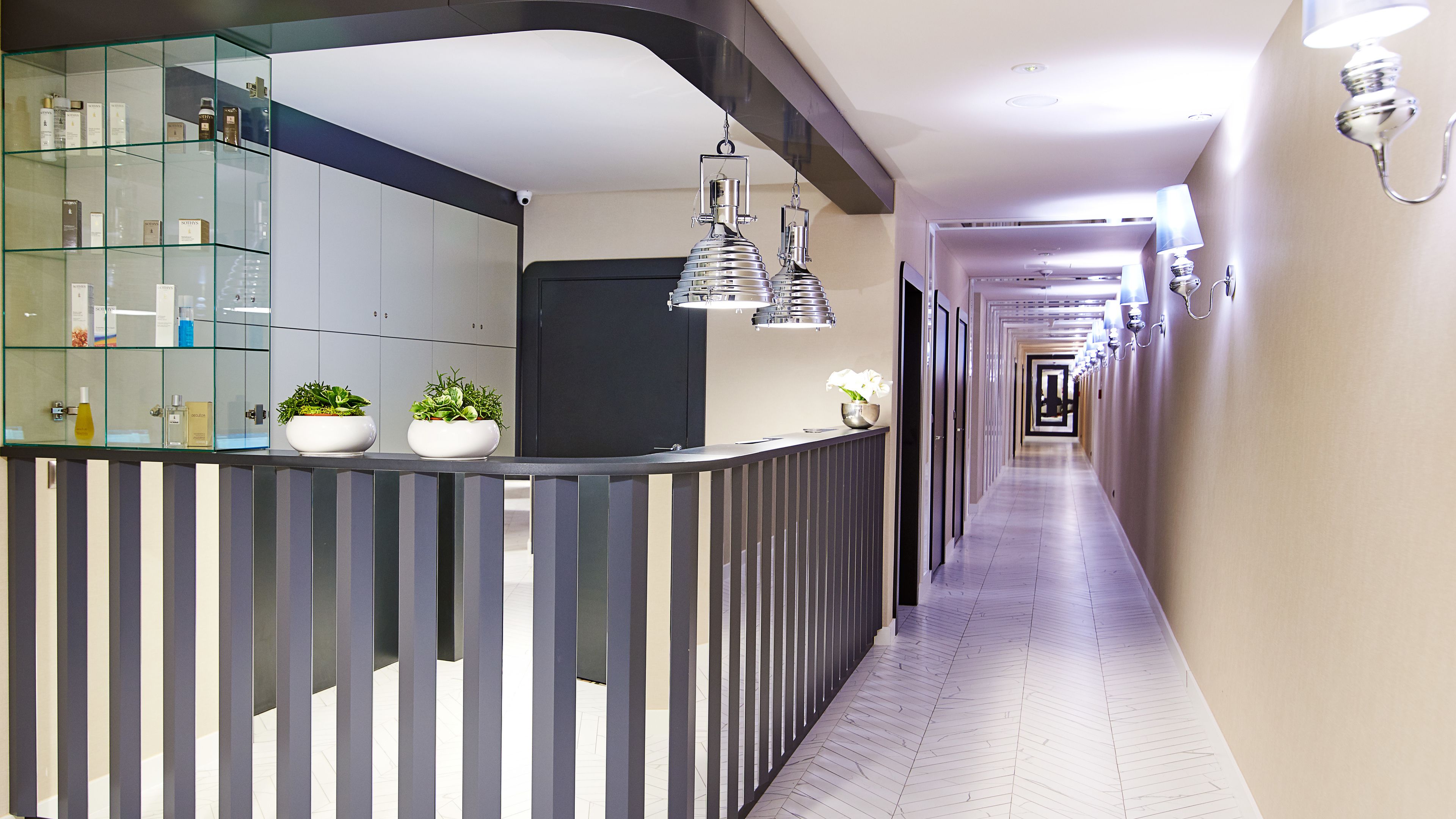 A modern hotel lobby with a reception desk, glass display cabinets, potted plants, and a long corridor with wall sconces.