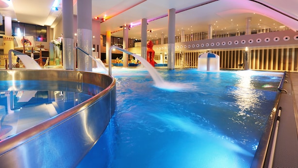 Ein großer, überdachter Schwimmbereich mit mehreren Bahnen und einer Rutsche, umgeben von hohen Decken und moderner Architektur.
