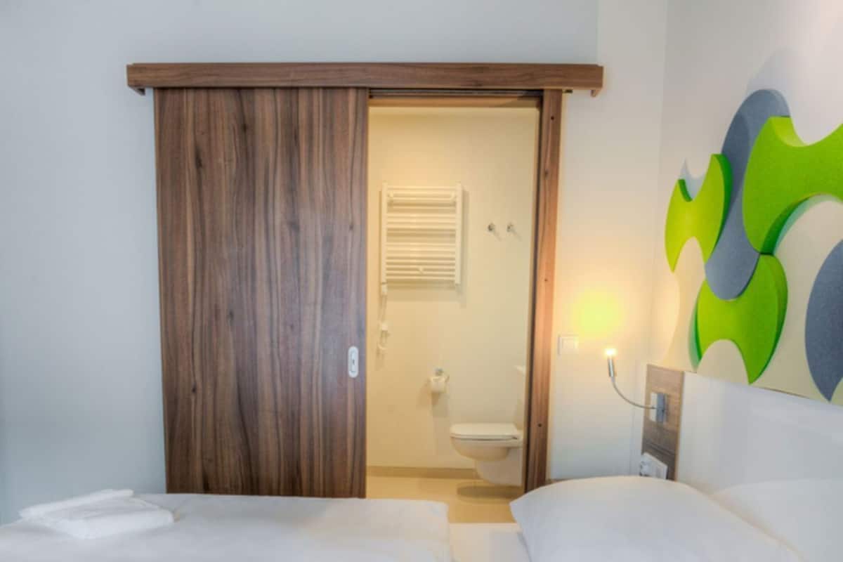 Ein Hotelzimmer mit zwei Betten, einem Holzschrank, einem Badezimmer mit Toilette und Dusche sowie einer Wand mit abstrakten Kunstwerken.