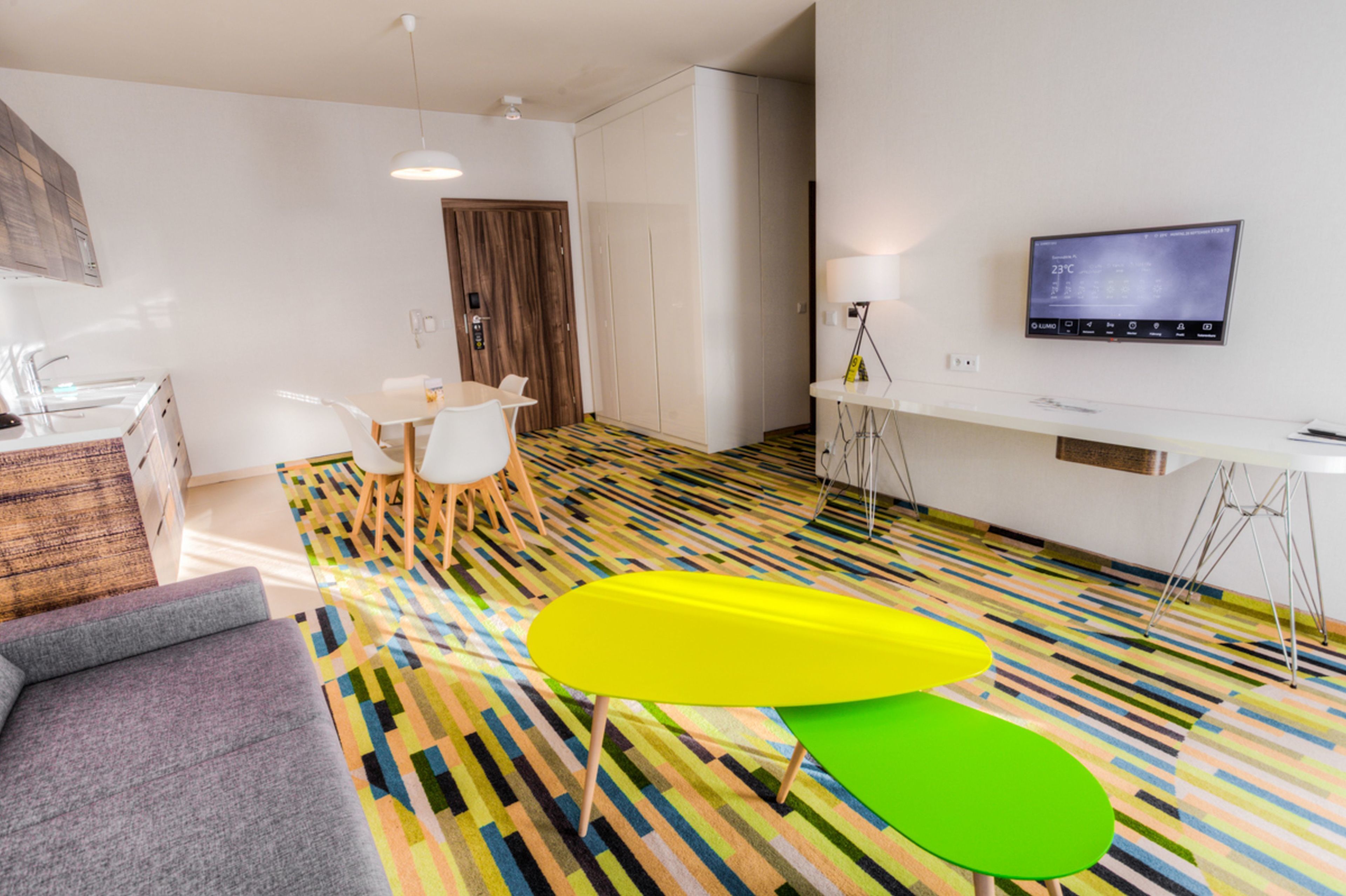 A modern hotel room with a colorful striped floor, a sofa, a dining table with chairs, a TV mounted on the wall, and a wardrobe.