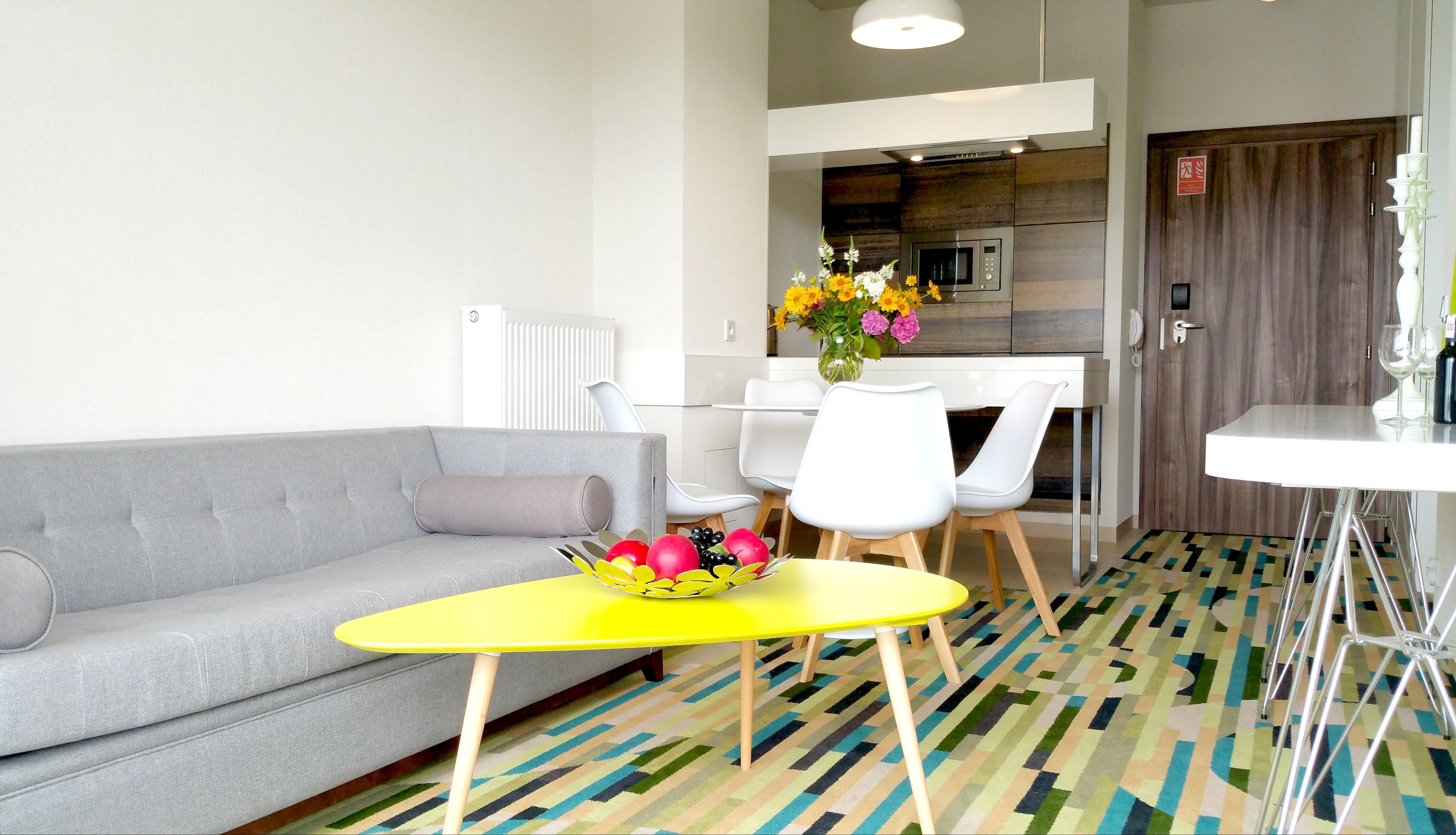A modern hotel room with a grey sofa, a yellow table with fruit, white chairs, a kitchen area, and a patterned floor.