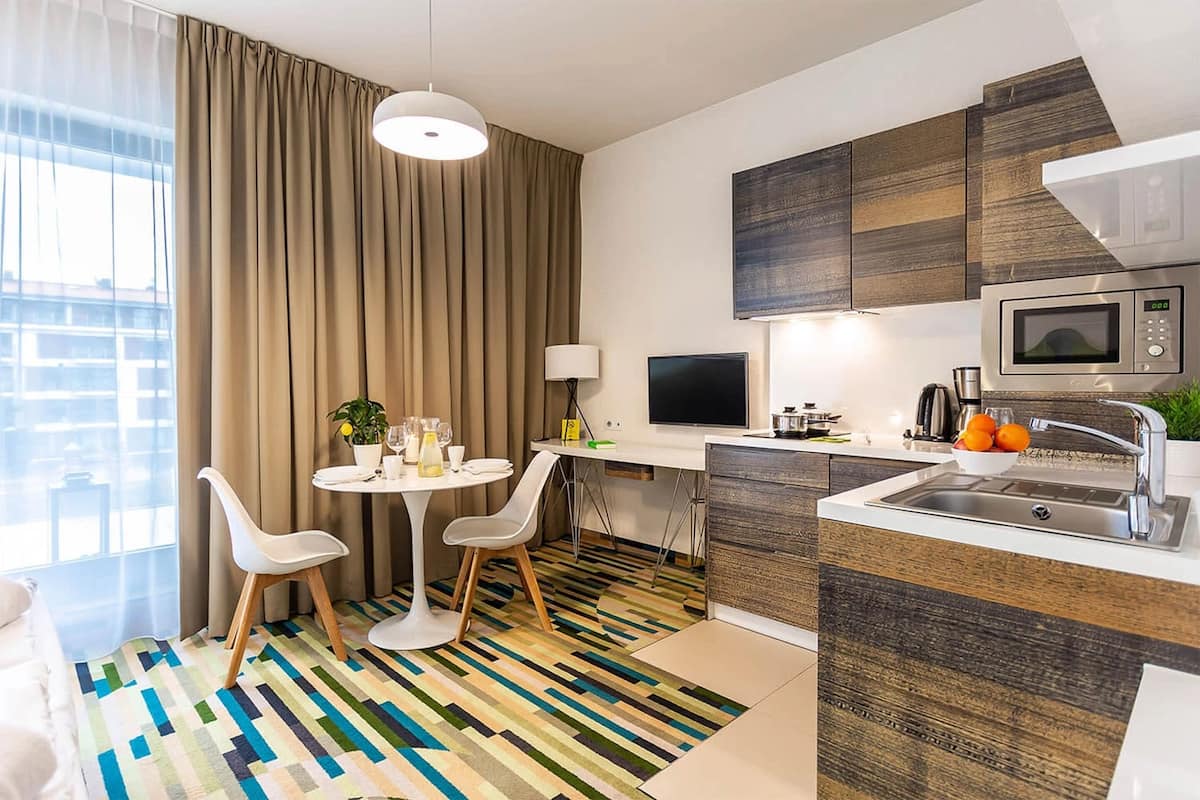 Ein modernes Hotelzimmer mit Essbereich, einer Küche inklusive Mikrowelle und einem großen Fenster mit Vorhängen.