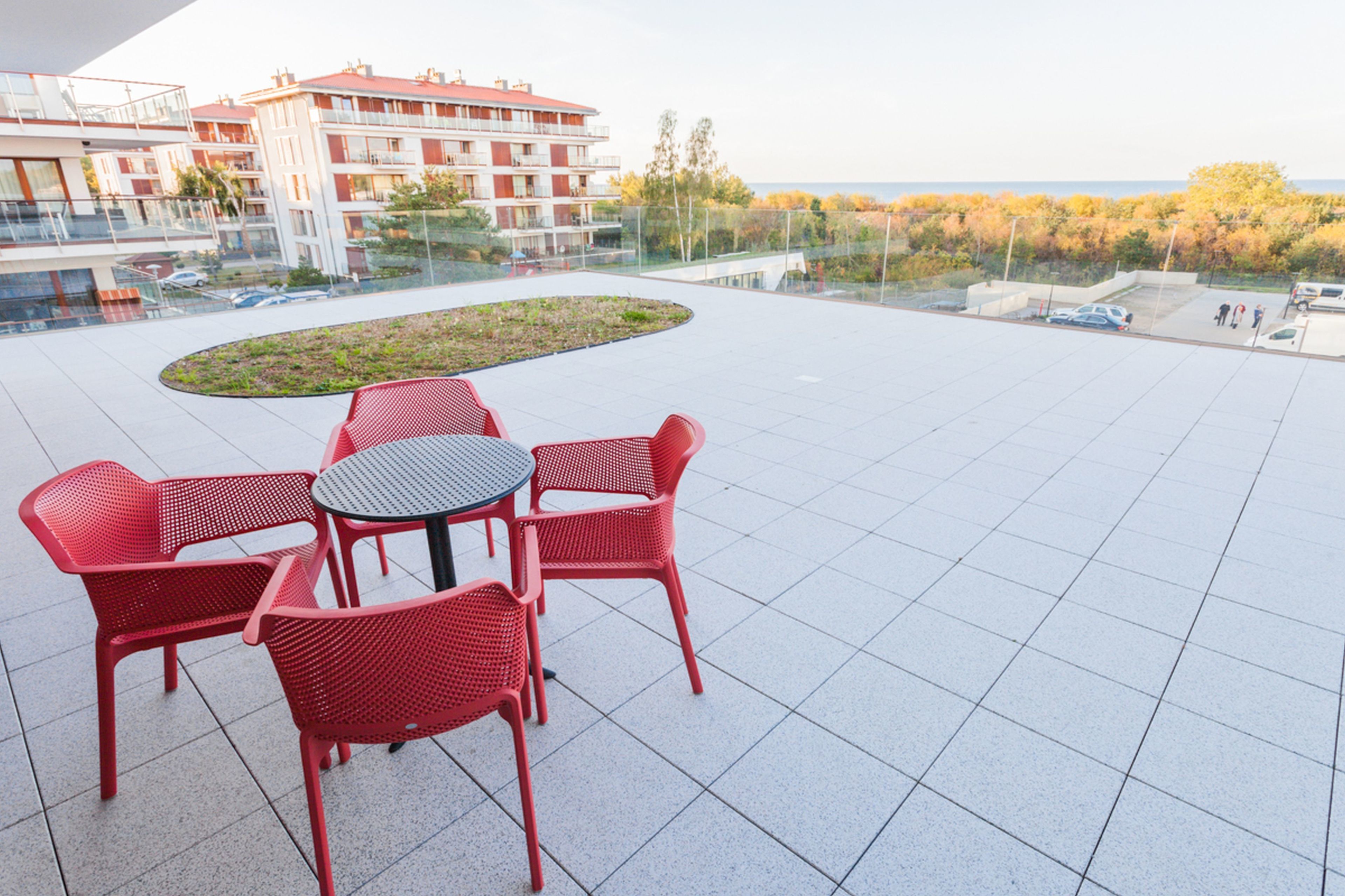 A rooftop terrace with red chairs and a table, overlooking a landscaped area and a body of water.