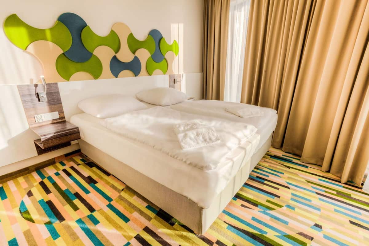 Ein Hotelzimmer mit einem Bett, einem Nachttisch, einer Wandlampe, einem farbenfrohen Streifen Teppich und einem Fenster mit Vorhängen.