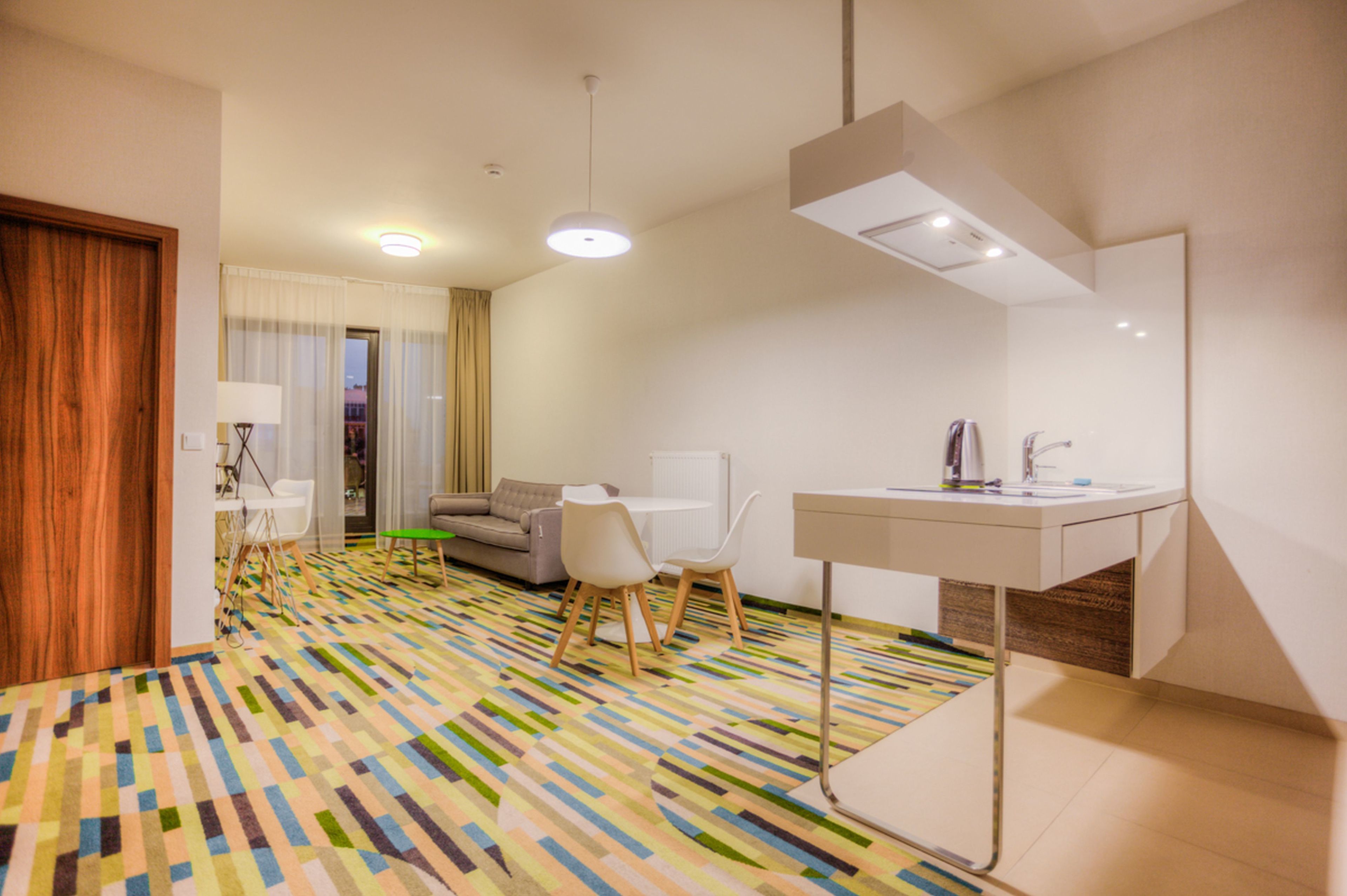 Ein modernes Hotelzimmer mit einem farbenfrohen geometrischen Teppich, einem weißen Schreibtisch mit Spiegel, einer Couch und einer Essecke mit Stühlen.