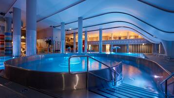 Una piscina interior moderna con un jacuzzi, rodeada de un espacio amplio con techo curvo y grandes pilares.