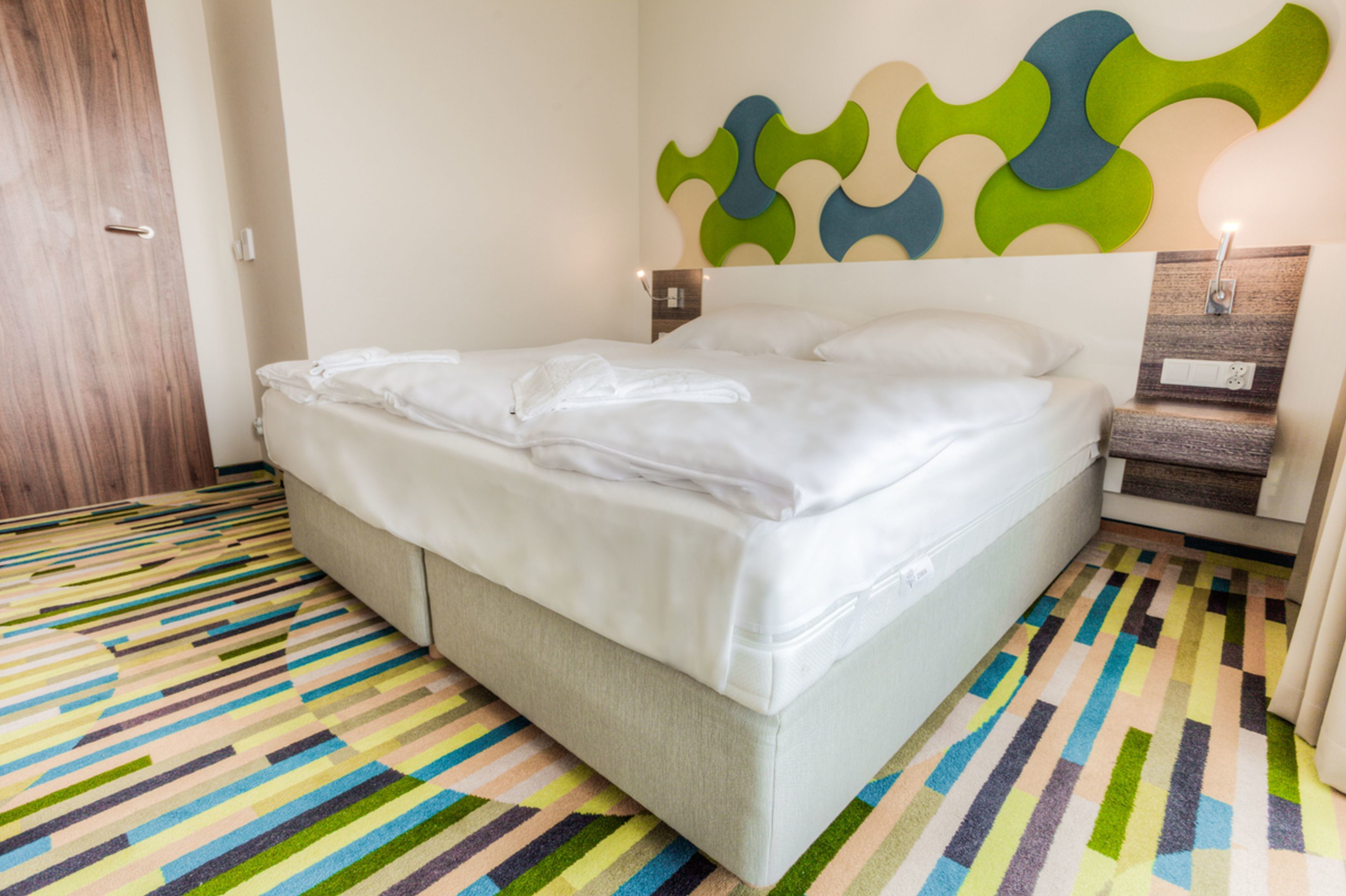 A hotel room with a large bed, a colorful striped carpet, a wooden door, and a wall with a geometric pattern.