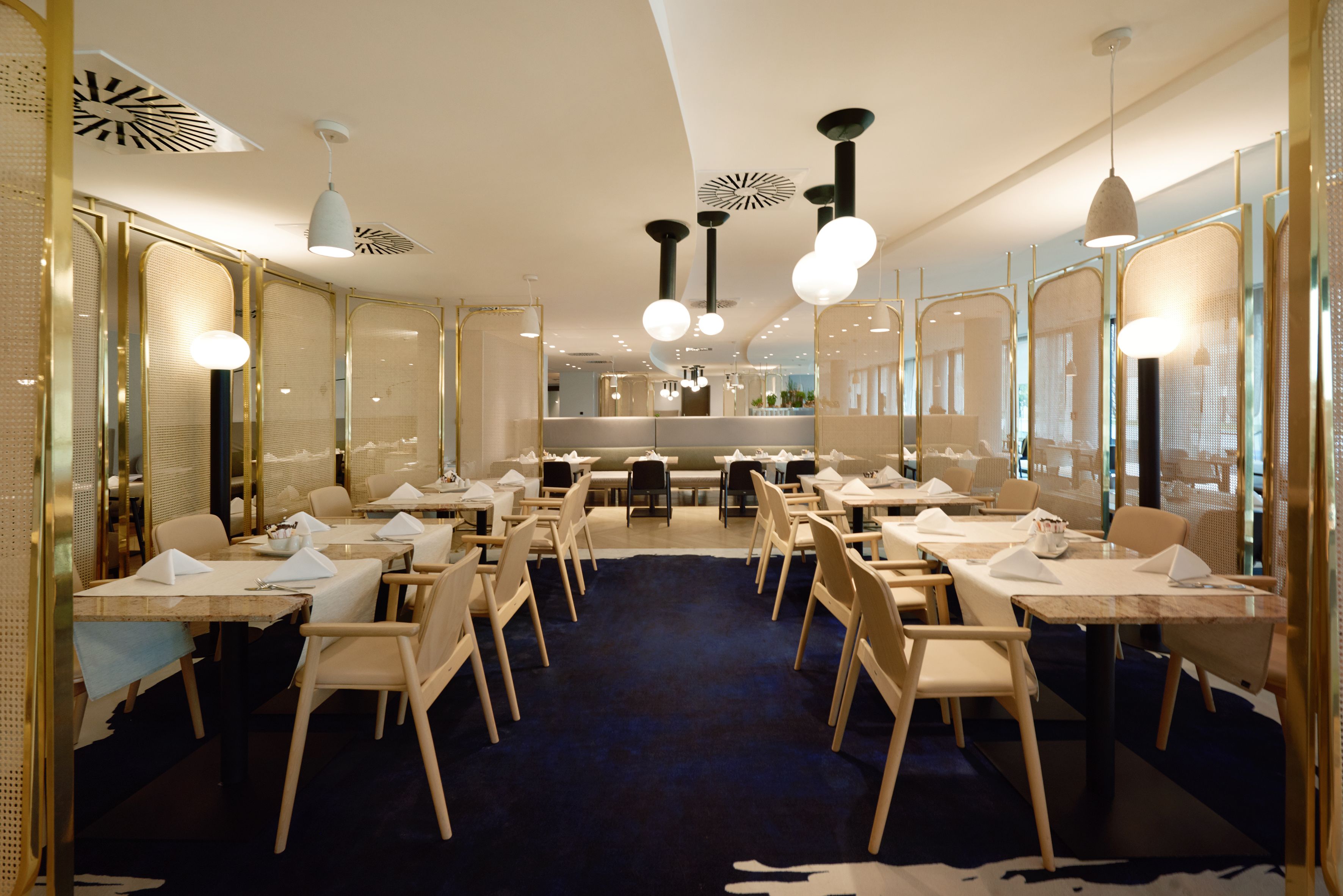 A modern dining area with multiple tables and chairs, a dark blue carpet, and contemporary lighting fixtures.