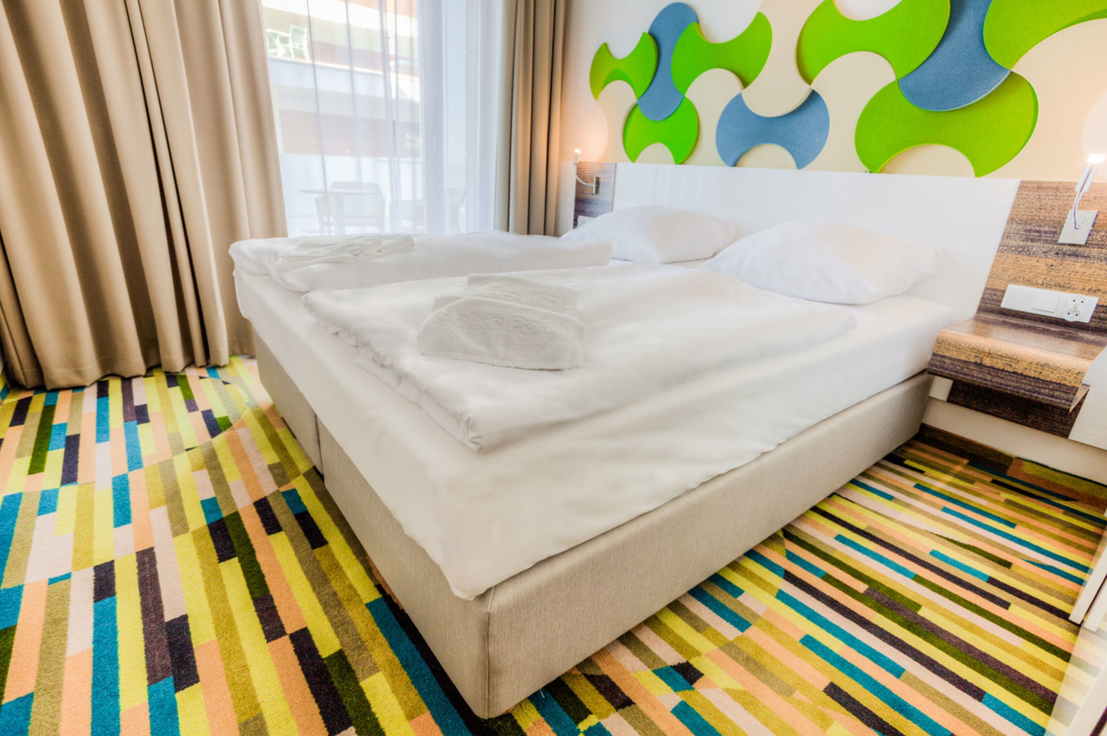 A hotel room with a neatly made bed, a colorful striped carpet, and a modern wall design.