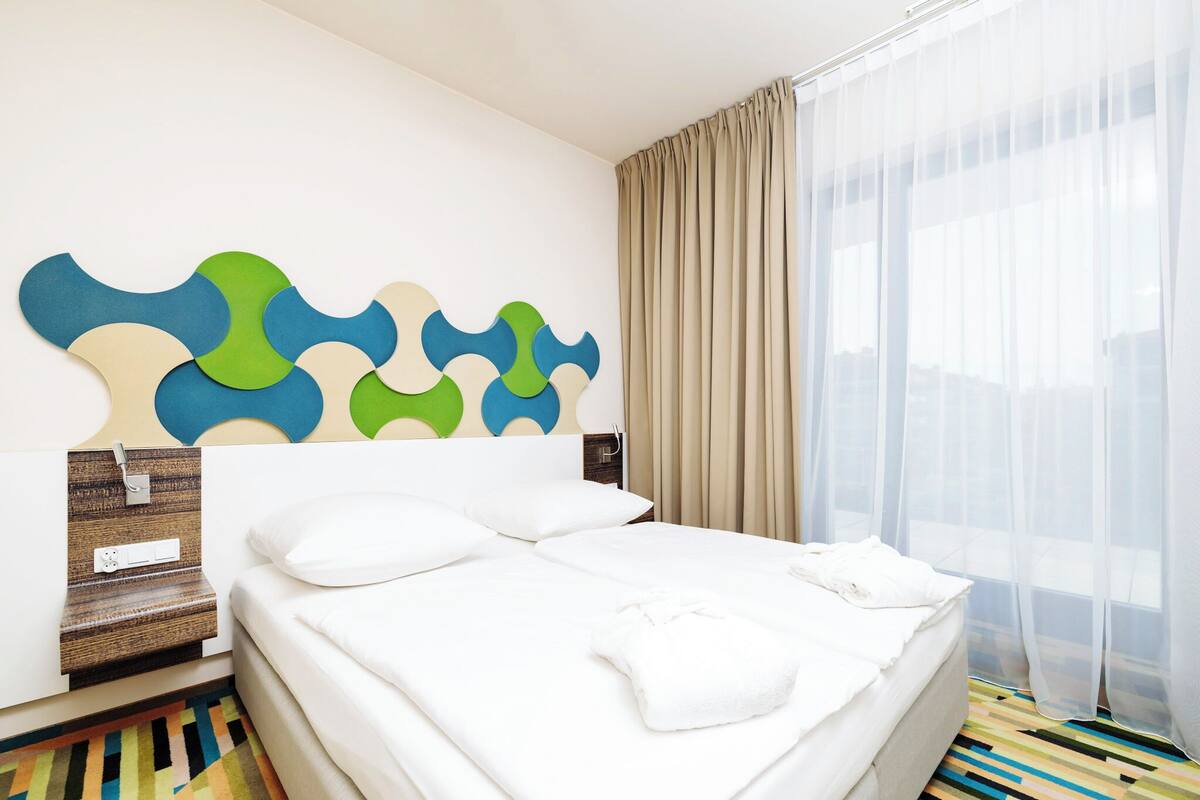 Ein Hotelzimmer mit einem großen Bett, einem an der Wand befestigten Fernseher, einem Nachttisch mit Lampe und einem Fenster mit durchsichtigen Vorhängen.