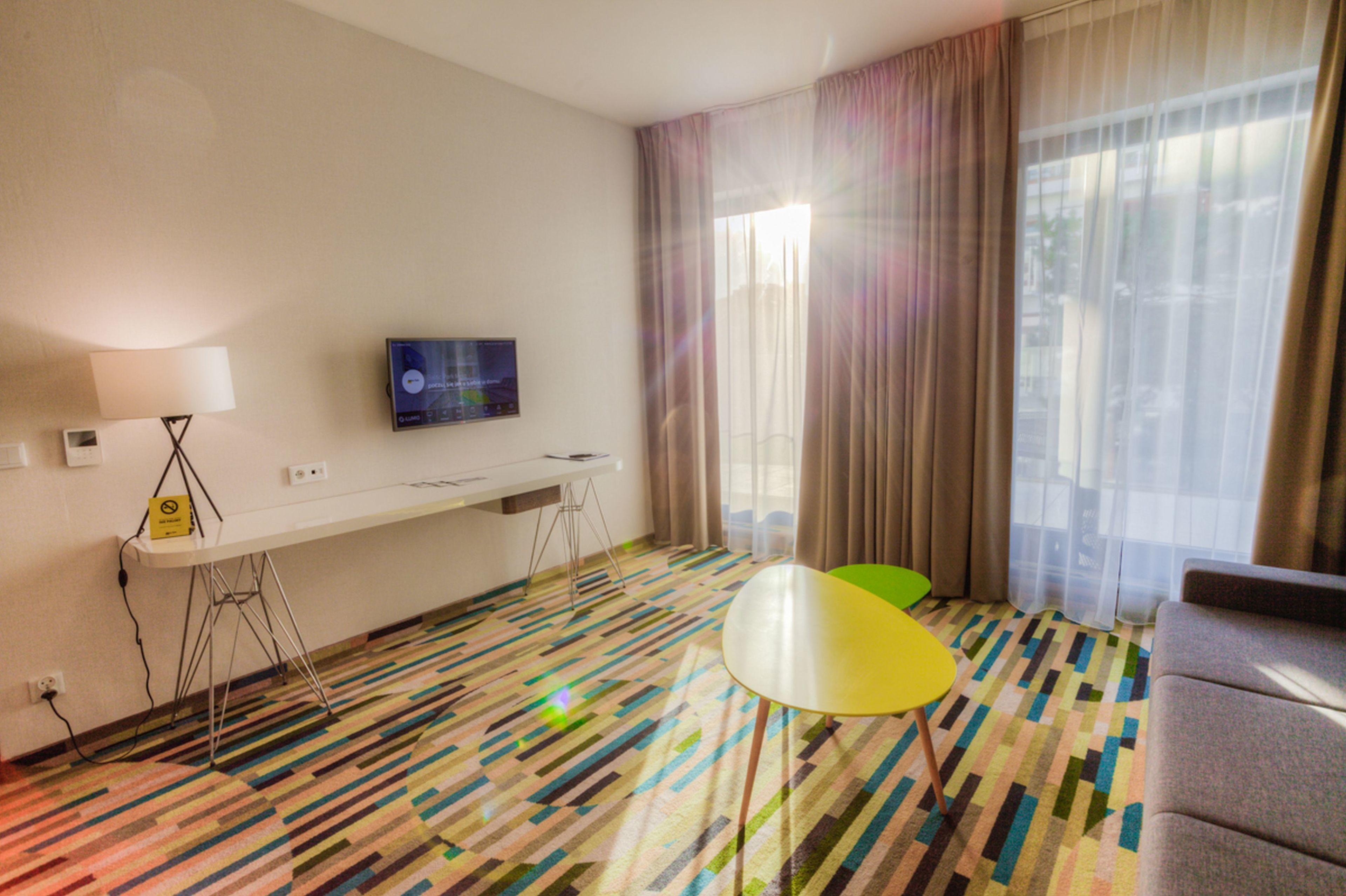 A hotel room with a long white table, a small yellow chair, a sofa, a TV on the wall, and a large window with sheer curtains.