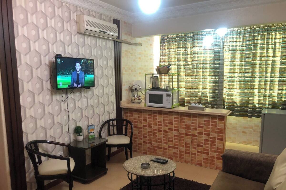 A living room with a TV, two chairs, a small table, a sofa, a kitchen area with a microwave, and a window with curtains.