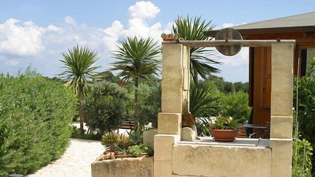 Une pergola en pierre avec une table et des chaises, entourée d’une végétation luxuriante et de palmiers.