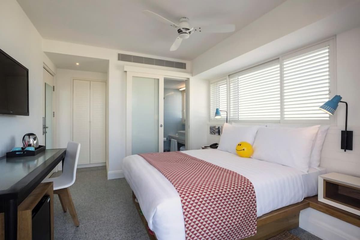 A bedroom with a bed, a desk, a chair, a TV, and a window with blinds.