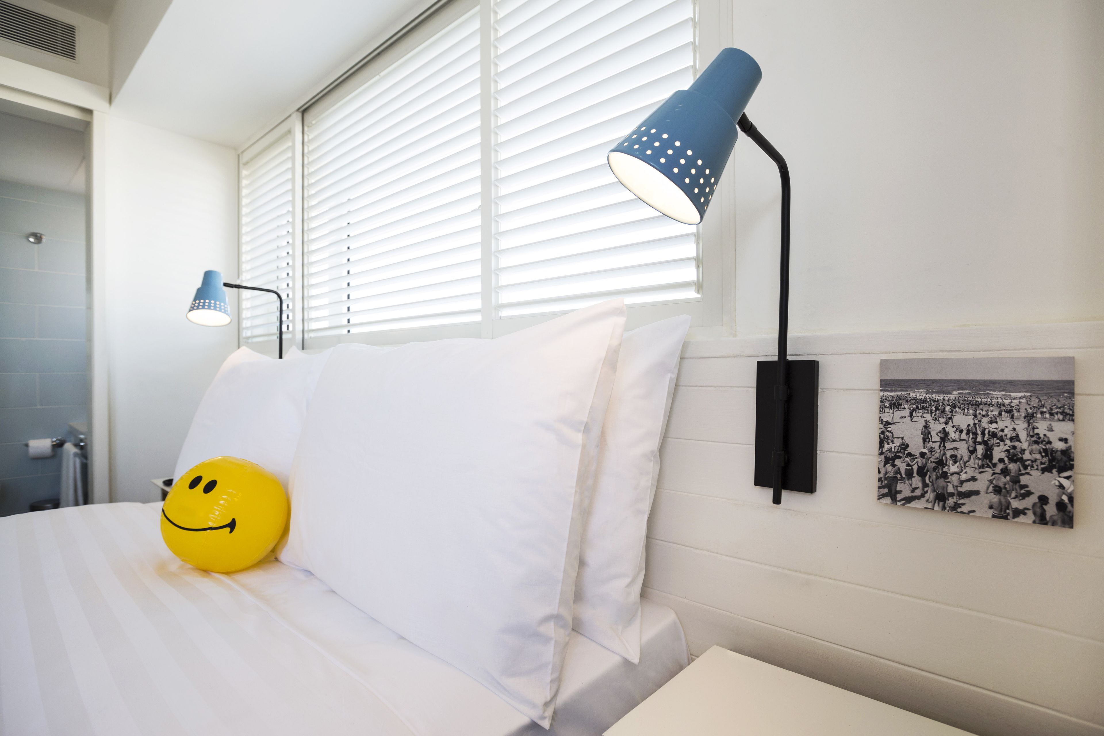 A hotel room with a bed, a smiley face pillow, a wall-mounted lamp, and a beach photo.