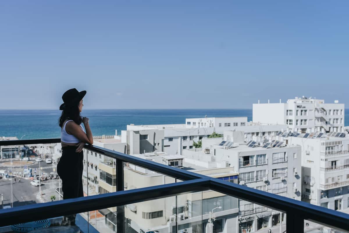 Una persona de pie en un balcón con vistas a un paisaje urbano, edificios y el mar.