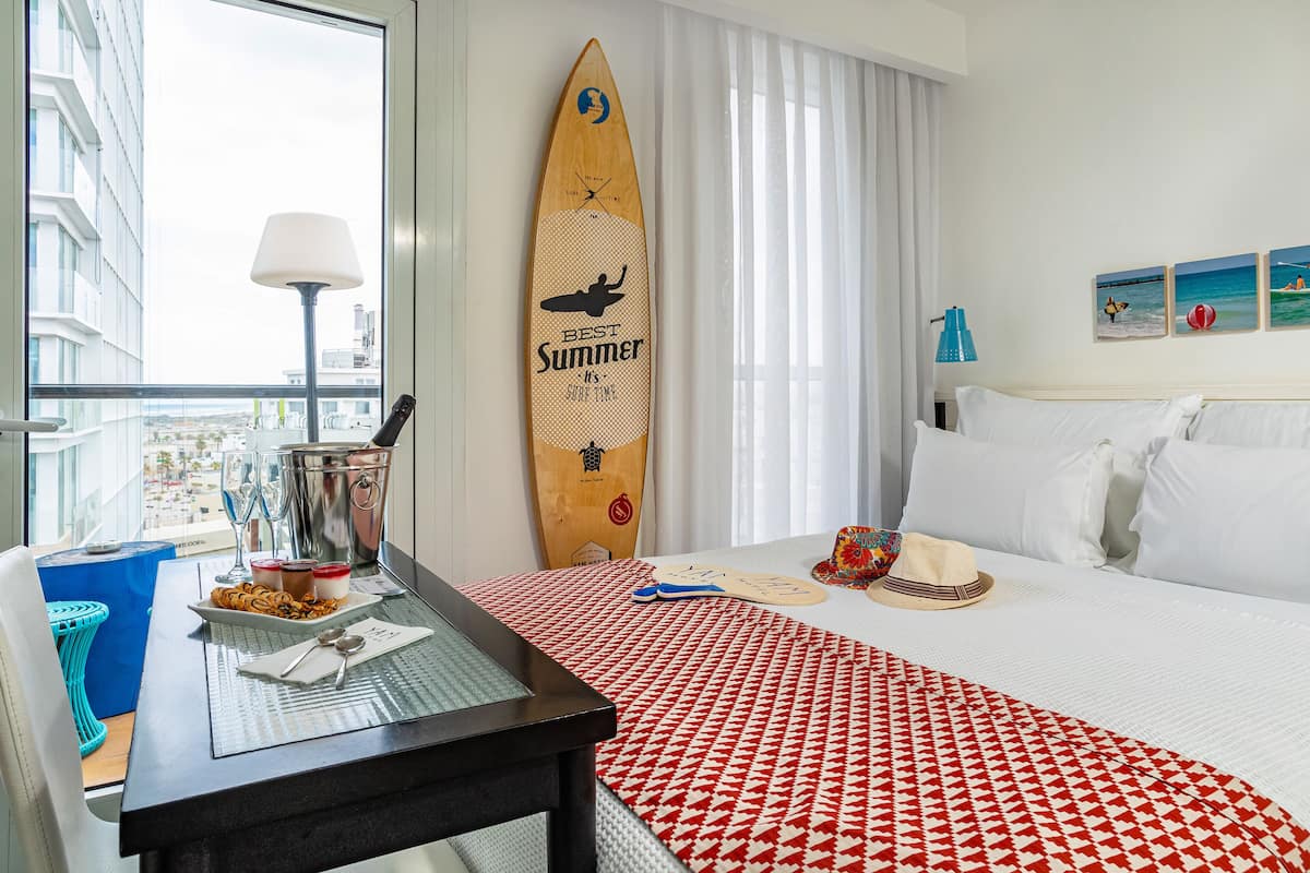 Habitación de hotel con cama, una mesa de comedor con desayuno, una tabla de surf y vistas a la ciudad.