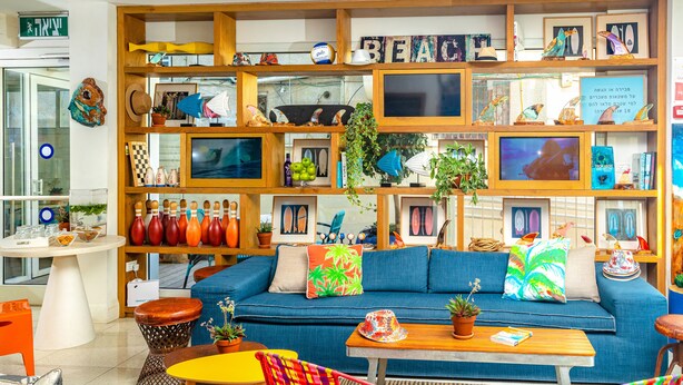 Una habitación con un sofá azul, estanterías de madera con diversos objetos, una mesita y sillas coloridas.