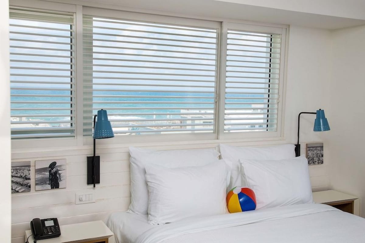 A hotel room with a bed, a nightstand with a phone, and a window with blinds.
