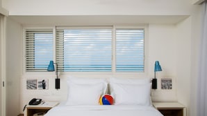 Habitación de hotel con cama, dos mesitas de noche, lámpara, pelota de playa y ventana con persianas.