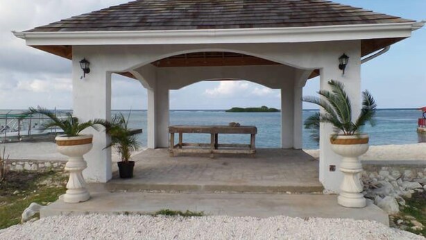 A gazebo with a bench, surrounded by white pillars and potted plants, overlooking a beach and ocean. - The Royal Kensington Villa (Montego Bay)