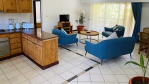 Una cocina moderna con armarios de madera y una isla central, un espacio de estar con sofás azules y una mesa de centro, y un ventanal con cortinas.