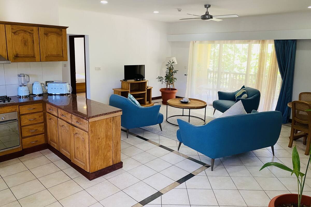 A modern kitchen with wooden cabinets and a central island, a living area with blue sofas and a coffee table, and a large window with curtains.