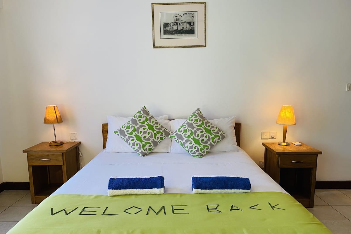 A neatly made bed with a green bedspread and pillows, two bedside lamps, and a framed picture on the wall.